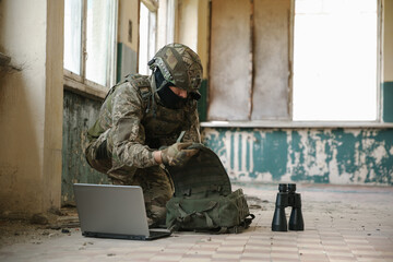 Military mission. Soldier in uniform using laptop and binoculars inside abandoned building