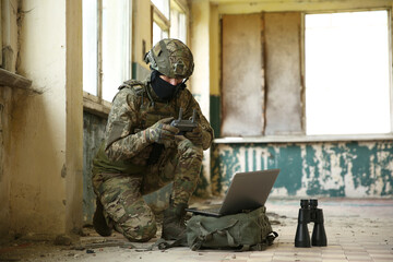 Military mission. Soldier in uniform using laptop and binoculars inside abandoned building