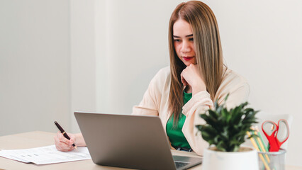A woman is sitting at a desk with a laptop and a pen. She is looking at the laptop screen and she...