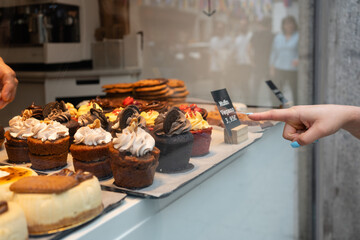 Woman pointing finger to vegan muffin in pastry shop window. Variety of delicious cupcakes are displayed in a bakery window, with a woman pointing at a vegan muffin