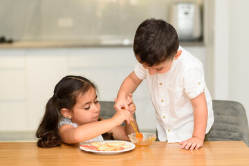 Fototapeta premium Little boy is leaning over a kitchen table, helping his sister dip apple slices in a small bowl of honey. He is holding honey dipper as she reaches to dip. They are both sitting at the table.
