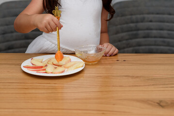 Young girl in a white dress sits at a wooden table, dipping slices of apple into a bowl of honey with a bee-shaped dipper.