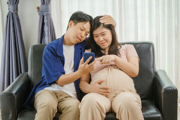 An Asian husband sits on a sofa, gently holding his pregnant wife's stomach. He lovingly cares for...