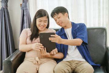 An Asian husband sits on a sofa, gently holding his pregnant wife's stomach. He lovingly cares for...