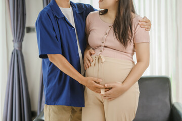 An Asian husband sits on a sofa, gently holding his pregnant wife's stomach. He lovingly cares for...