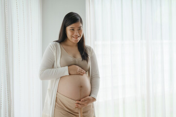 An Asian woman stands while pregnant with her first child, placing her hand on her stomach. She is...