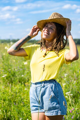 Woman wearing yellow shirt and straw hat standing in sunny field with green grass and wildflowers.