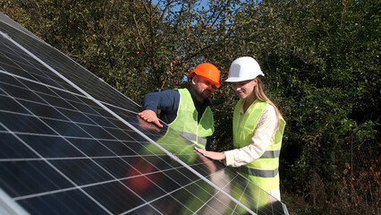A man and a woman in hard hats discuss work tasks near solar panels on the street. Female...