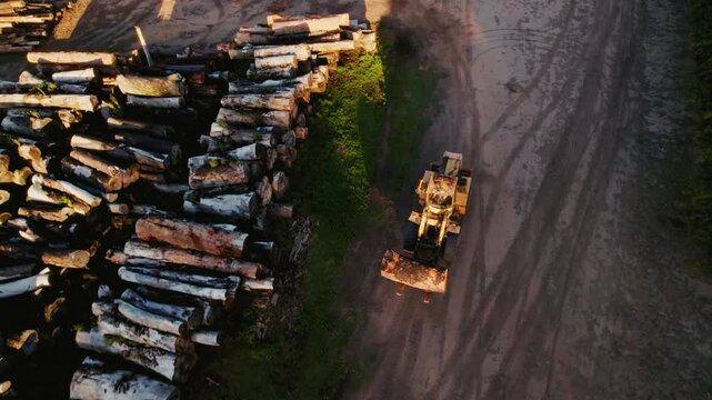An aerial view of a yellow loader parked on a dirt road next to a large pile of cut logs in a rural setting.