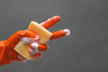 Hand in orange rubber glove holding washing sponge and shows like of success sign with soapy foam. Isolated on gray background for cleaning and washing concept.