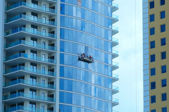 Workers on an outside suspended scaffold platform cleaning a skyscraper building facade with glass windows and balconies . - Powered by Adobe