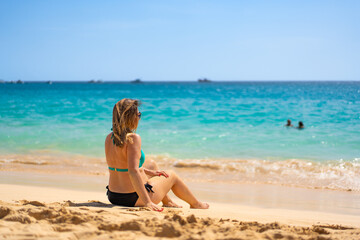 Beach vacation. Happy young woman in bikini sitting on sandy beach by the sea on beautiful sunny day. Positive emotion
