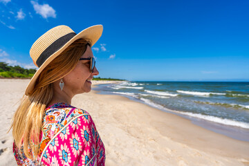 Beach holidays on Baltic Sea. Smiling young plus size woman wearing fashionable multicolored pink dress with hat standing on sunny sandy beach on windy day. Side portrait
