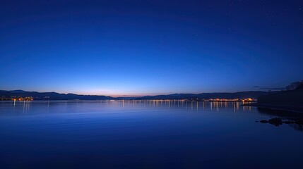 Calm water reflecting city lights at blue hour under starry sky
