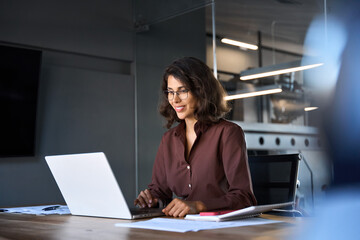 Smiling 30s latin hispanic middle-aged business woman working on laptop computer in modern office. Indian young businesswoman professional employee using pc doing online banking analysing at workplace