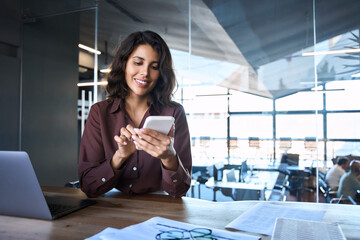 Young Indian business woman manager ceo using cell phone mobile app laptop. Smiling Latin Hispanic mature female businesswoman holding smartphone in office trading, reading, working online. Copy space