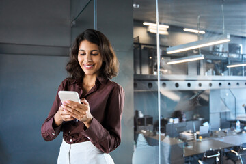 Happy young Latin Hispanic business woman holding mobile cell phone, watching app for work. Smiling middle eastern entrepreneur businesswoman holding smartphone cellphone standing in modern office