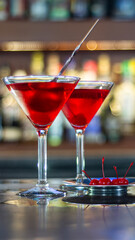Two martini glasses brimming with the delicious Cosmopolitan cocktail, garnished with fresh cherries. In the background, a bar is filled with bottles of fine liquor, ready to be enjoyed! 