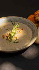 A delicious, creamy mushroom soup, garnished with crunchy almonds and aromatic rosemary, served in a beautiful bowl. In the background, you can see pieces of freshly baked bread and a black tablecloth