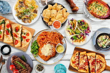 Table scene of various delicious foods. Above view on a white wood background. Pastas, flatbreads, tacos, steak and restaurant theme dishes.