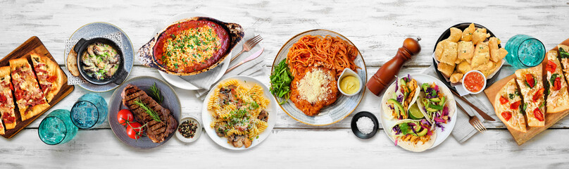 Table scene of various delicious foods. Top view on a white wood banner background. Pastas, flatbreads, tacos, steak and restaurant theme dishes.