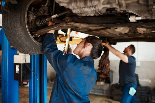 Mechanic, woman and man under car in workshop for auto repair, service and inspection for safety. Engineering, partnership and teamwork for vehicle maintenance with tools, parts or industry at garage