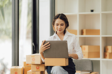 Asian businesswoman in white shirt uses laptop to do online shopping at her store. A sales woman prepares a package to send to a customer.