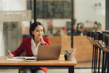 Focused and Determined: A young businesswoman in a red blazer sits at her desk, engrossed in her...