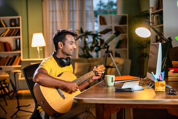 Asian indian young musician plays or learns guitar online using computer or smartphone