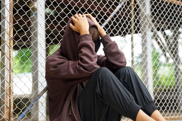 A young Asian male wearing a hood sits with his face in his hands and his head in his hands, his back resting against a wire mesh fence in a juvenile detention center.
