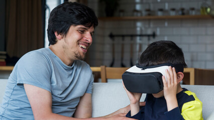 A father and son spending quality time at home. Boy wearing VR headset, experiencing immersive technology, while father watches and smiles. Modern family interaction and bonding.