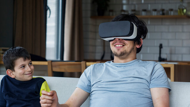 A father and son enjoy bonding over virtual reality in a modern living room. The father wears a VR headset, interacting with the virtual world, while the son smiles and sharing in the excitement.