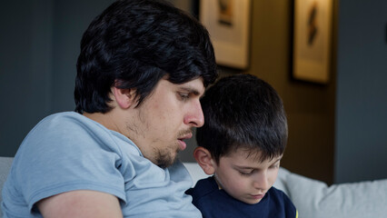 A close-up shot of a father and son sitting on a sofa, engaging with a tablet. Moment of family interaction, technology in fostering learning and connection within the home environment.	