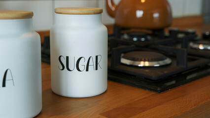 Close-up shot of stylish white ceramic jars labeled sugar with wooden lids on a wooden kitchen counter, creating an organized and chic storage solution for kitchen essentials