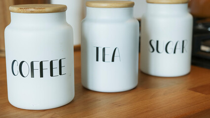 Close-up shot of stylish white ceramic jars labeled coffee, tea and sugar with wooden lids on a...