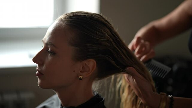 Closeup of unrecognizable hairdresser brushing wet long hair with comb after washing. Close-up face of blonde female client getting hair dressed in beauty salon. Concept of fashion hairstyling.