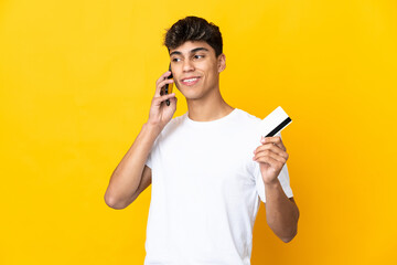 Young man over isolated yellow background keeping a conversation with the mobile phone and holding a credit card