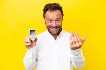 Middle age man holding a engagement ring isolated on yellow background inviting to come with hand. Happy that you came