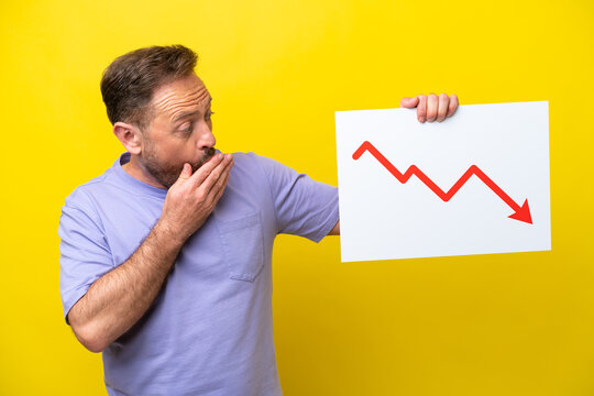 Middle age caucasian man isolated on yellow background holding a sign with a decreasing statistics arrow symbol with surprised expression