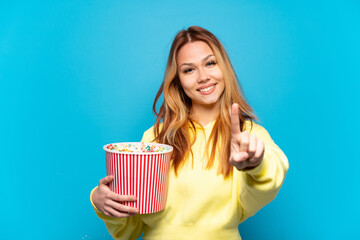 Teenager girl holding popcorns over isolated blue background showing and lifting a finger