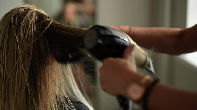 Closeup cropped shot of unrecognizable female hairstylist drying and straightening long blonde client hair with comb and hairdryer in beauty salon. Concept of professional fashion hairstyling.
