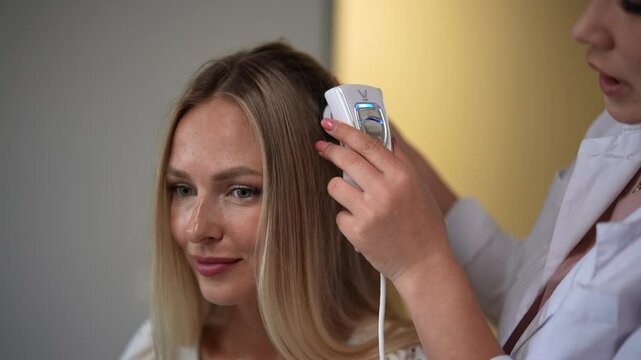 Blonde female client visiting trichologist in dermatological clinic. Close-up hands of unrecognizable dermatologist making diagnosis of structure and condition of hair with special device trichoscope.