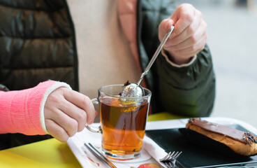 woman with a pink plaster cast on her right arm sits in a cafe and drinks tea with eclair, active...