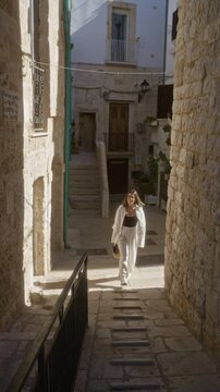 A young hispanic woman walks through the charming stone streets of polignano a mare in puglia, italy, bathed in sunlight and surrounded by historic architecture.