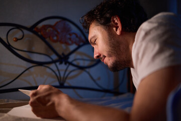 A man is peacefully reading a book in bed in a dimly lit bedroom, covered with cozy blankets