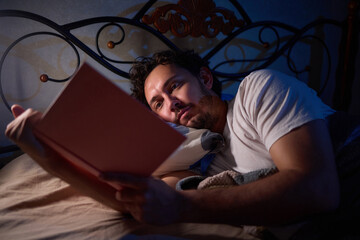 A man is peacefully reading a book in bed in a dimly lit bedroom, covered with cozy blankets