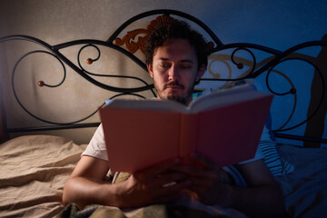 A man is peacefully reading a book in bed in a dimly lit bedroom, covered with cozy blankets