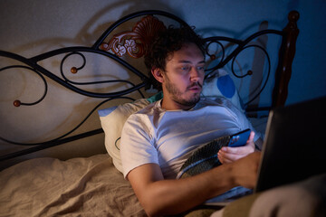 A bearded man using smartphone in dimly lit bedroom, lying comfortably in bed late at night