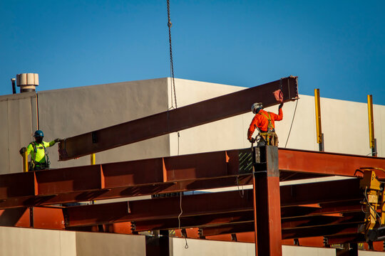 Two construction workers directing placement of a large steel beam that is atttached to a crane