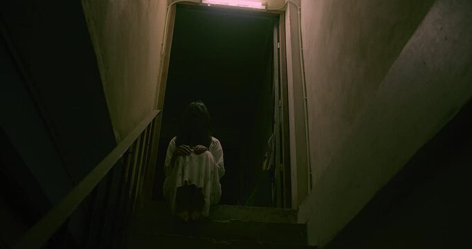 Horror scene of a mysterious Scary Asian ghost woman creepy have hair covering the face sitting on staircase at abandoned house with background dark scene movie at night, festival Halloween concept